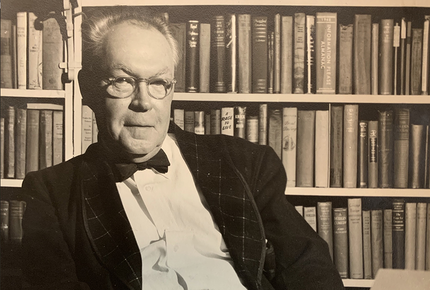 Author sits in front of a bookcase. He wears a bow tie and jacket with decorative lapels. 